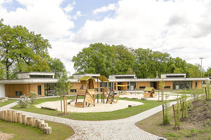 Kindergarten-Spielgeräte von eibe – große Spielanlage aus Holz auf einer sandigen Fläche zwischen mehreren Gebäuden eines Kindergartens. 